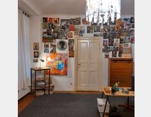 Ein Zimmer mit weiß gestrichenen Wänden, dekoriert mit vielen kleinen Bildern und Collagen rund um eine cremefarbene Tür. Es gibt ein Regal mit gestapelten Büchern, einen kleinen Holztisch mit Lampe sowie einen Schreibtisch mit Schreibutensilien. Ein auffälliger Kronleuchter hängt von der Decke.