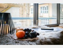 Fensterbank in einem Innenraum mit großer Fensterfront; darauf stehen eine schwarze Laterne mit Kerze, ein Holzbrett mit Obst sowie ein Laptop auf einem flauschigen Fell. Durch das Fenster sind gegenüberliegende Gebäude mit Ziegel- und Putzfassaden und mehreren Fenstern zu sehen.