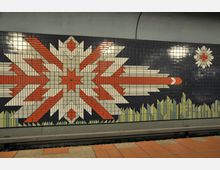 Wandgestaltung in einer U-Bahn-Station mit einem Mosaik aus farbigen Kacheln, das ein geometrisches Muster in Rot, Weiß, Grün und Schwarz zeigt, abstrakt an eine Blume oder einen Stern erinnernd. Im unteren Bereich sind stilisierte Pflanzen dargestellt, der Boden besteht aus terrakottafarbenen Fliesen.