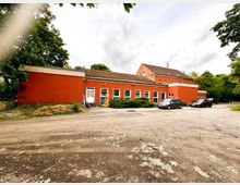 Flaches, langgezogenes rotes Gebäude mit vielen rechteckigen Fenstern und mehreren Eingängen, dahinter ein höherer Gebäudeteil mit Satteldach. Davor liegt ein großer, unbefestigter Parkplatz mit einigen abgestellten Autos, umgeben von Bäumen und Grünflächen.