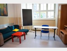 Ein einfacher Innenraum mit einer Sitzgruppe aus einem türkisfarbenen Sofa, einem roten Beistelltisch und einem gelben Kissen. Rechts stehen ein weißer Tisch mit einem blauen Stuhl, ein großes Fenster, ein schwarzes Display auf einem Ständer sowie ein Holzschrank.