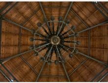 Blick nach oben in eine achteckige Holzdachkonstruktion mit sichtbaren Metallstreben. In der Mitte hängt ein großer, sternförmiger Kronleuchter mit vielen radial angeordneten Armen.