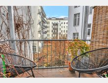 Blick von einem Balkon mit Holzfußboden und Metallgeländer, umgeben von Kletterpflanzen und Bambusmatten, auf einen Innenhof mit herbstlichem Laub und umliegenden mehrstöckigen Wohnhäusern mit weißen Fassaden und Balkonen.