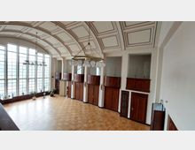 Innenraum eines historischen Versammlungsraums mit Holzvertäfelungen, hohen Fenstern mit Sprossen und einem gewölbten, dekorativ gestalteten Deckenbereich. Der Raum verfügt über eine hängende Lampe mit mehreren kugelförmigen Leuchten und einen Parkettboden.