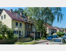 Reihenhäuser mit roten Ziegeldächern an einer ruhigen Wohnstraße, umgeben von Bäumen und kleinen Gärten mit Holzzäunen; geparkte Autos stehen entlang der Straße.