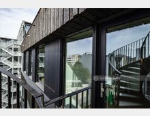 Fassadendetail eines modernen Gebäudes mit einer Mischung aus Holz- und Glaselementen, großen Fenstern und einer außenliegenden Wendeltreppe. Im Hintergrund sind weitere Gebäude sowie Bäume in einer urbanen Umgebung sichtbar.