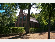 Backsteinbau mit Giebeldach und großen Fenstern, umgeben von einer Hecke und hohen Bäumen in einer bewaldeten Umgebung. Der Boden ist teilweise mit Laub bedeckt, und die Szene liegt im Schatten der Bäume.