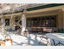 Überdachte Terrasse eines Hauses mit mehreren Tischen und Stühlen aus Holz und Metall. Die Fassade ist mit großen vergitterten Fenstern versehen, und Kletterpflanzen ranken an den Pfeilern empor; der Boden ist mit hellem Kies bedeckt.