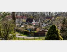 Wohnsiedlung mit rot gedeckten Häusern, kleinen Gärten und umgebendem Grün. Im Vordergrund ein Weg, Zäune und vereinzelt Bäume, im Hintergrund dichter Baumbestand.