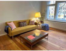 Wohnzimmer mit einem gelben Sofa, dekoriert mit Kissen und einer blauen Decke. Vor dem Sofa steht ein flacher Couchtisch mit Büchern, daneben eine Tischlampe auf einem kleinen Beistelltisch. Große Fenster lassen Tageslicht herein, und der Raum hat einen Holzboden sowie eine dekorative Zimmerpflanze in der Ecke.