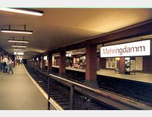Unterirdischer U-Bahnhof mit zwei gegenüberliegenden Bahnsteigen, bezeichnet mit „Mehringdamm“. Die Decke ist mit rechteckigen Leuchtstofflampen beleuchtet, und die Wände sowie Säulen bestehen aus dunklem Material. Schienen verlaufen zwischen den Bahnsteigen.
