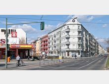 Straßenszene in einer Stadt mit einem Eckgebäude im klassizistischen Stil, das weiße Fassaden und Balkone besitzt, sowie mehreren Geschäften im Erdgeschoss. Links sind bunte Wohnhäuser und ein kleiner Laden mit rotem Schild zu sehen, während rechts die mehrspurige Straße verläuft.