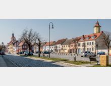 Straßenszene einer Altstadt mit gepflasterten Gehwegen und niedrigeren Gebäuden mit roten, grauen und beigefarbenen Dächern. Im Hintergrund befinden sich ein Kirchturm und ein runder Wachturm mit spitzem Dach; kahle Bäume und Straßenlaternen säumen den Weg.