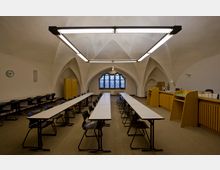 Ein historisch anmutender Seminarraum mit gewölbter Decke und spitzbogigen Arkaden. Der Raum enthält lange Tische mit schwarzen Stühlen, eine Holztheke auf der rechten Seite und ein großes Fenster mit verzierten Gittern im Hintergrund.