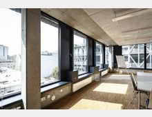 Heller Konferenzraum mit großen, bodentiefen Fenstern, die den Blick auf benachbarte moderne Gebäude mit Glasfassaden ermöglichen. Der Raum hat Holzboden, Sichtbetonwände und eine Decke mit integrierter Beleuchtung; im Vordergrund sind ein Tisch, Stühle und ein Flipchart zu sehen.