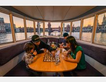 Eine Innenansicht eines Bootes, mit großen Fenstern, die einen Blick auf die Oberbaumbrücke und umliegende Gebäude am Flussufer bei Sonnenuntergang bieten. Am Tisch in der Mitte des Raumes sitzen vier Personen und spielen ein Brettspiel, während auf dem Tisch Getränke und Snacks stehen.