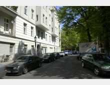 Wohnstraße mit mehrstöckigem Wohngebäude mit heller Fassade auf der linken Seite und parkenden Autos entlang der Straße. Rechts säumen üppige grüne Bäume den Straßenrand, die Schatten auf die Fahrbahn werfen.