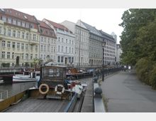 Historische Häuserfassaden mit verzierten Fassaden und roten Dächern säumen einen Kanal, an dem mehrere Boote festgemacht sind. Rechts verläuft ein Gehweg mit Laternen und Grünflächen.