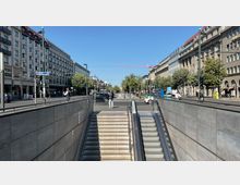 Treppen- und Rolltreppenabgang einer U-Bahn-Station an einer belebten Stadtstraße, flankiert von mehrstöckigen Gebäuden mit klassischen Fassaden und Geschäften im Erdgeschoss, Bäumen am Straßenrand und einer roten Baukran-Silhouette im Hintergrund.