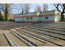 Freiluftbühne mit terrassenförmig angeordneten Sitzreihen aus Metall und einer kleinen, rechteckigen Bühne vor einem Gebäude aus hellgrünen und ziegelroten Elementen. Im Hintergrund sind Bäume und ein Zaun zu sehen.