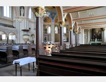 Innenraum einer Kirche mit hölzernen Kirchenbänken, Säulen mit verzierten Kapitellen und einer bemalten Decke. Im Hintergrund befinden sich ein Altar, ein großes Gemälde und ein kunstvoll gestalteter Kanzelbereich.