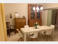 Esszimmer mit einem weißen Tisch, umgeben von weißen Stühlen mit Holzbeinen. Im Hintergrund stehen zwei massive Holzschränke, links ein niedriger Schrank und rechts ein großer Vitrinenschrank. Eine Pendelleuchte und ein Blumenstrauß auf dem Tisch dienen als Dekoration.