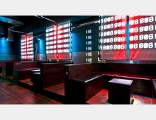 Innenansicht einer modernen Lounge mit Holzbänken und niedrigen Tischen. Die Wände sind mit leuchtenden LED-Panels in rot und weiß gestaltet, die geometrische Muster zeigen, und der Boden besteht aus Holzplanken.