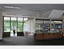 Eine einfache Bahnhofshalle mit einem kleinen Verkaufskiosk, der Süßwaren, Zeitungen und Reisebedarf anbietet. Im Hintergrund ist ein großer verglaster Ausgang zu sehen, durch den man ins Freie mit Bäumen blickt; eine Person verlässt gerade das Gebäude.