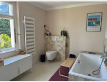 Modernes Badezimmer mit beiger Farbgestaltung, freistehender Badewanne, Wandtoilette und einem großem Fenster, das den Blick ins Grüne ermöglicht. Ein Handtuchheizkörper, ein kleiner Schrank, Fliesenakzente und eine aufgeräumte Einrichtung prägen die minimalistische Gestaltung.