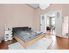 Schlafzimmer mit einem Holzbett und grauer Bettwäsche auf einem weißen Teppich. Im Hintergrund führen weiße Doppeltüren in ein angrenzendes Esszimmer mit Holzboden, Esstisch und Stühlen.