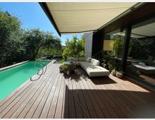 Moderne Terrasse mit Holzdeck, angrenzendem rechteckigen Pool und einer weißen Outdoor-Sofaecke. Im Hintergrund befinden sich dichte grüne Bäume, während eine Markise teilweise Schatten spendet.