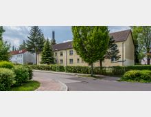 Wohngebiet mit einer gepflasterten Straße, zweistöckigen Mehrfamilienhäusern mit gelber Putzfassade und Satteldächern, umgeben von Bäumen, Sträuchern und gepflegten Hecken. Straßenlaternen und ein Straßenschild sind sichtbar.