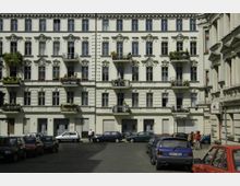 Fassade eines historischen Wohngebäudes mit verzierten Fenstern, Balkonen und Blumenkästen, umgeben von parkenden Autos auf einem gepflasterten Platz; ein Straßenschild mit „Wilhelm-August-Straße“ ist sichtbar.