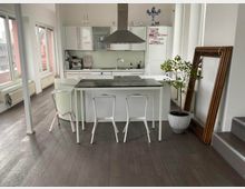 Bright modern kitchen with white cabinets and large windows, featuring a central island with a dark countertop and white chairs beneath a stainless-steel range hood. A potted indoor tree and a large framed mirror lean against the wall near a short set of stairs.
