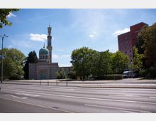 Eine Moschee mit einer gestreiften Fassade, einer grünen Kuppel und einem Minarett, gelegen an einer Straße mit mehreren Fahrspuren. Im Hintergrund sind Bäume und ein mehrstöckiges rotes Gebäude sichtbar.