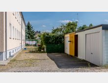 Ein Hinterhof mit gepflastertem Weg, flankiert von einem mehrstöckigen, beigefarbenen Wohngebäude und Garagen mit gestrichenen Türen. Im Hintergrund sind Wäscheständer, ein grüner Zaun, Bäume und ein Teil eines Wohnhauses sichtbar.