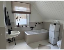 Modernes Badezimmer mit schräger Decke und einem weißen Fliesendesign. Es verfügt über eine Badewanne mit eingehängter Dusche, ein kleines Waschbecken, einen rechteckigen Spiegel, einen offenen Aufbewahrungsschrank mit drei Fächern und ein Fenster mit halbtransparentem Rollo; auf der Fensterbank stehen Dekorationen wie Trockenblumen und kleine Pflanzen.