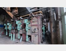 Industriehalle mit historischen Maschinen, darunter grüne, vertikale Kolbenapparate vor einer Ziegelwand. Rohrleitungen, Anzeigen und Ventile dominieren die Szene, begleitet von metallischen Plattformen und Geländern im Hintergrund.