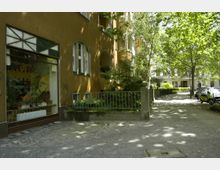 Straßenszene mit einem mehrstöckigen Wohngebäude in gelber Putzfassade, großen Fenstern und einem begrünten Eingangsbereich mit Geländer. Daneben stehen Gehwege, Bäume, parkende Autos und weitere Wohnhäuser im Hintergrund.