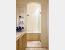 Modernes Badezimmer mit einer freistehenden Badewanne aus beigem Stein, umgeben von cremefarbenen Wandfliesen. Links ein Waschtisch mit Holzverkleidung und daneben Regalnischen mit dekorativen Pflegeprodukten; rechts eine Glastür zur Duschzone.