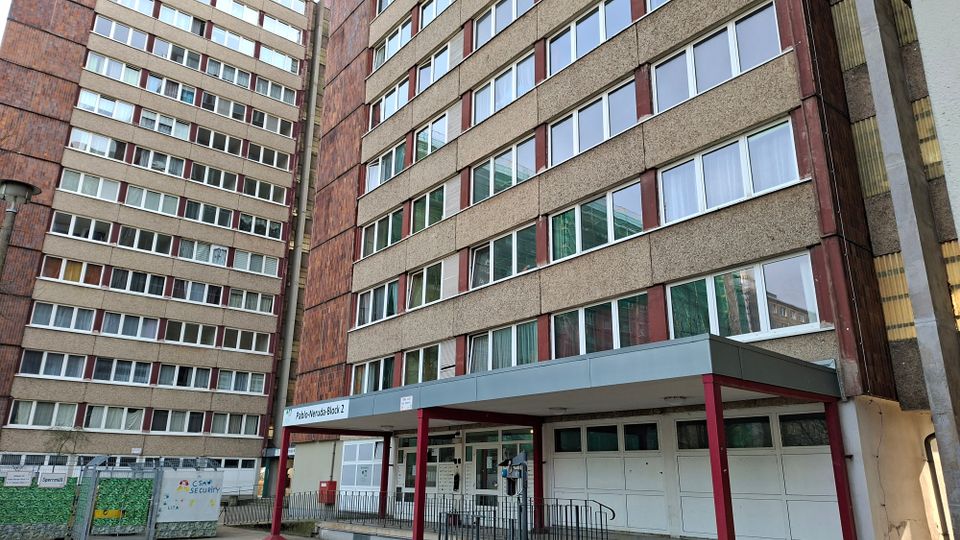 Eingangsbereich eines hohen Wohnblock-Komplexes mit rauer Betonfassade und vielen horizontalen Fensterreihen; ein flaches Vordach auf roten Stützen markiert den Zugang. Links stehen weitere mehrstöckige Gebäudeflügel, im Vordergrund sind Zaun, Garagentore und befestigte Wege zu sehen.