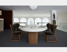 Ein moderner Essbereich mit einem ovalen weißen Tisch auf einem hölzernen Sockel, umgeben von acht gepolsterten Stühlen mit dunklen Stoffbezügen und Holzrahmen. Im Hintergrund befinden sich bogenförmige Fenster mit weißen Vorhängen und eine schwarze Skulptur auf einem Sockel.