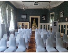 Ein eleganter Raum mit Reihen weiß bezogener Stühle, einem Holzboden und grün gemusterten Wänden. An den Wänden hängen Bilder und Vorhänge, ein antikes Regal ist sichtbar, und durch eine offene Tür im Hintergrund ist ein weiterer Raum erkennbar.