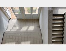 Treppenhaus mit gefliesten Stufen, das von oben fotografiert wurde. Im Hintergrund sind verglaste Türen und ein Außenbereich mit Steinplatten und etwas Grün zu sehen.