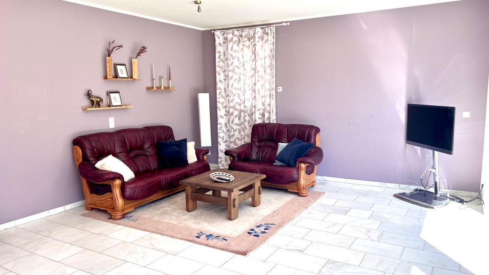 Wohnzimmer mit lilafarbenen Wänden, hellem Fliesenboden und einem großen Fenster mit gemusterten Vorhängen. Zwei bordeauxrote Ledersofas um einen niedrigen Holztisch auf einem Teppich, dazu Wandregale und ein Fernseher auf einem Standfuß.