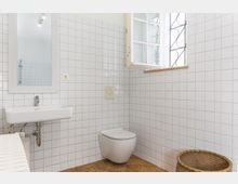 Minimalistisch eingerichtetes Badezimmer mit weißen Fliesen an den Wänden, einem hängenden Waschbecken mit Spiegel, einer Wandtoilette und einem kleinen geflochtenen Korb auf dem gefliesten Boden. Rechts befindet sich ein Fenster mit weißen Rahmen und dekorativem Gitter.