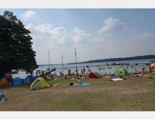Ein Strandbad an einem See mit Wiese im Vordergrund, auf der Zelte, Decken und Liegestühle verteilt sind. Im Hintergrund ist ein Badesteg mit Badenden sowie der See und eine bewaldete Uferlinie zu sehen.