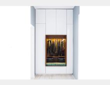 Ein moderner Einbauschrank mit weißen, glatten Oberfläche und einer zentralen offenen Nische aus Holz, die beleuchtet ist. In der Nische hängen Jacken an einer Kleiderstange, darunter befinden sich zwei schwarze Boxen und eine gepolsterte Sitzbank.