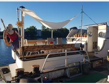 Ein Hausboot mit einer Terrasse auf dem Deck, dekoriert mit Pflanzen in Töpfen, einem Tisch mit Stühlen sowie einer Schaufensterpuppe mit Schwimmring. Im Hintergrund ist ein Fluss und eine Baumreihe unter klarem, blauem Himmel sichtbar.