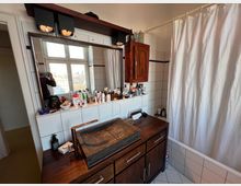 Badezimmer mit einem Waschtisch aus dunklem Holz und einem dunklen Steinausgussbecken, darüber ein großer Spiegel mit Lampen; links ein Fenster mit Stadtblick, rechts eine Badewanne mit weißem Duschvorhang. Wände sind weiß gefliest.